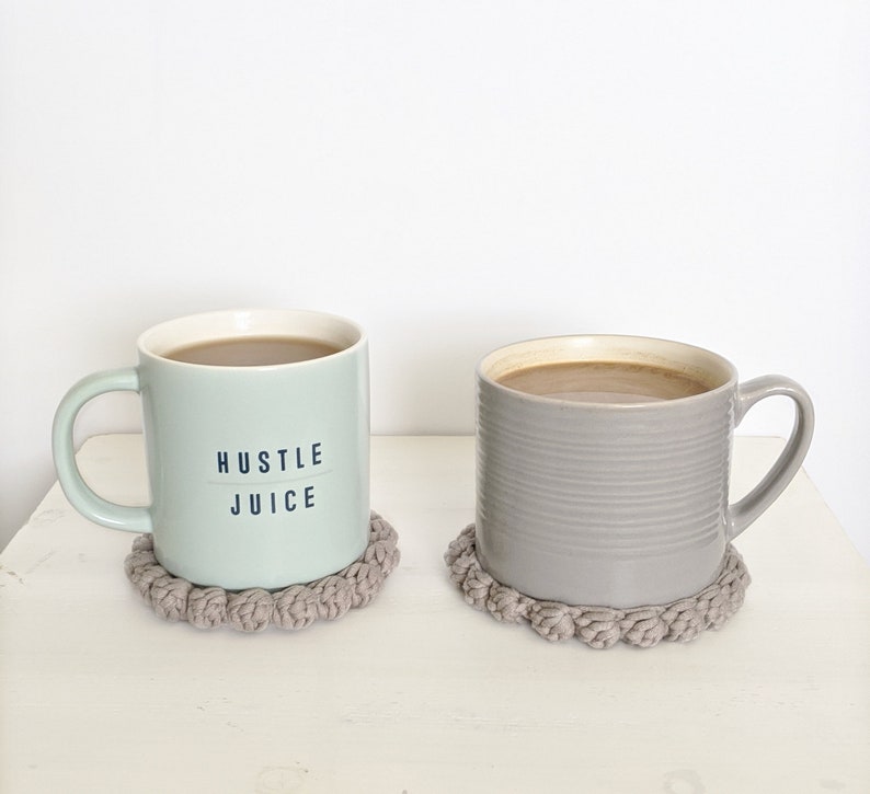 Puede incluir: Dos tazas con caf&eacute; o t&eacute; sobre posavasos de crochet grises. La taza de la izquierda es verde claro con el texto "Hustle Juice". La taza de la derecha es gris.