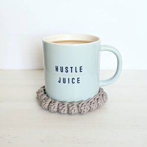 Puede incluir: Una taza de cer&aacute;mica azul claro con el texto "Hustle Juice" impreso en ella. La taza est&aacute; sobre un posavasos de crochet beige.
