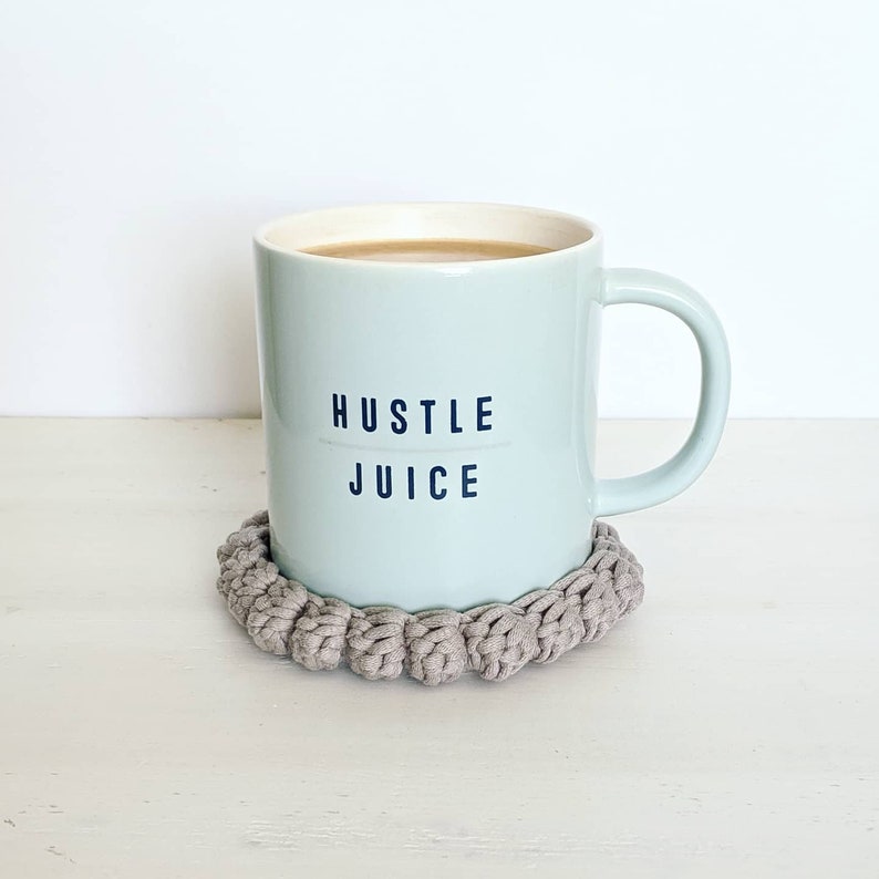 Puede incluir: Una taza de cer&aacute;mica azul claro con el texto "Hustle Juice" impreso en ella. La taza est&aacute; sobre un posavasos de crochet beige.