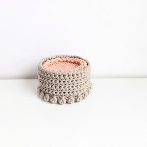 Puede incluir: Una cesta de crochet gris con un forro de crochet rosa. La cesta es redonda y tiene un dise&ntilde;o texturizado. La parte inferior de la cesta tiene una fila de pompones.