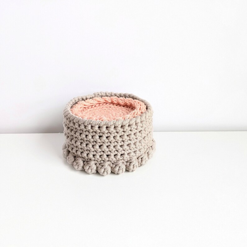 Puede incluir: Una cesta de crochet gris con un forro de crochet rosa. La cesta es redonda y tiene un dise&ntilde;o texturizado. La parte inferior de la cesta tiene una fila de pompones.