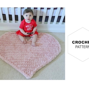 May include: A pink heart-shaped crochet rug with a baby sitting on it. The rug is made with a fluffy yarn and has a textured surface. The baby is wearing a red shirt with the words "Bee Mine" on it.