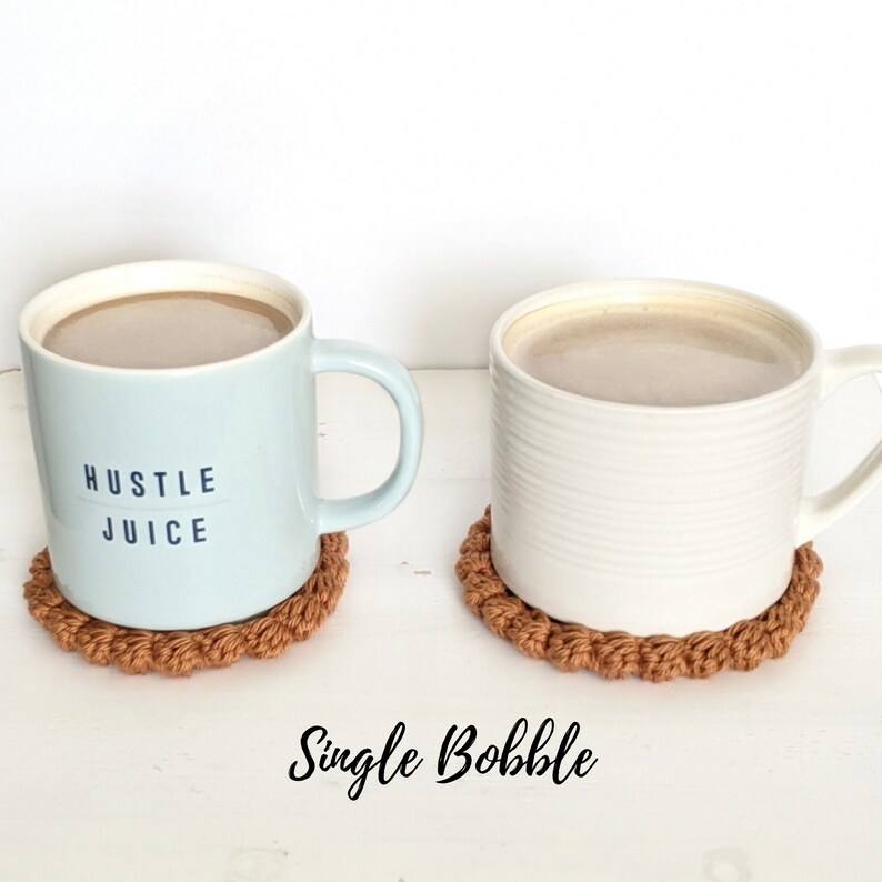 Puede incluir: Dos tazas de caf&eacute; sobre posavasos de crochet marrones. La taza de la izquierda es azul claro con el texto "Hustle Juice". La taza de la derecha es blanca con un dise&ntilde;o acanalado. Los posavasos est&aacute;n hechos con un punto bobble simple.