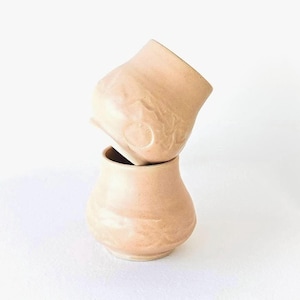 Könnte beinhalten: Zwei gestapelte, hellpfirsichfarbene Keramikvasen. Die obere Vase ist geneigt und gibt den Blick in das Innere der unteren Vase frei. Die Vasen haben eine strukturierte Oberfläche und eine abgerundete Form. Der Hintergrund ist weiß.