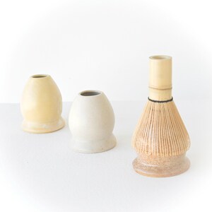 May include: Three items for preparing matcha tea. Two ceramic chawan rests in cream and yellow hues, and a bamboo chasen whisk. The chasen has a light brown base and a light brown whisk with a black band.
