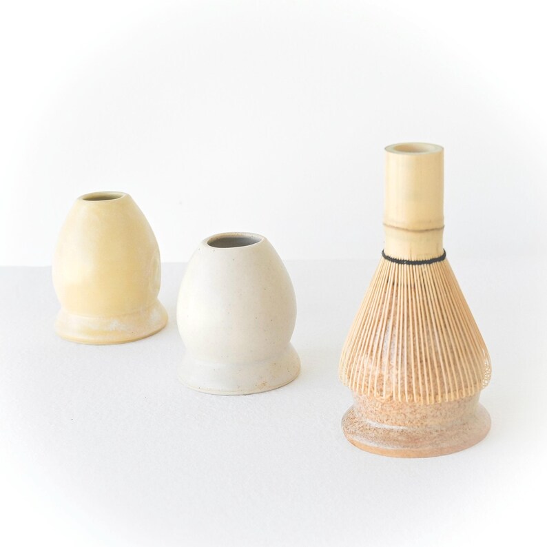 May include: Three items for preparing matcha tea. Two ceramic chawan rests in cream and yellow hues, and a bamboo chasen whisk. The chasen has a light brown base and a light brown whisk with a black band.