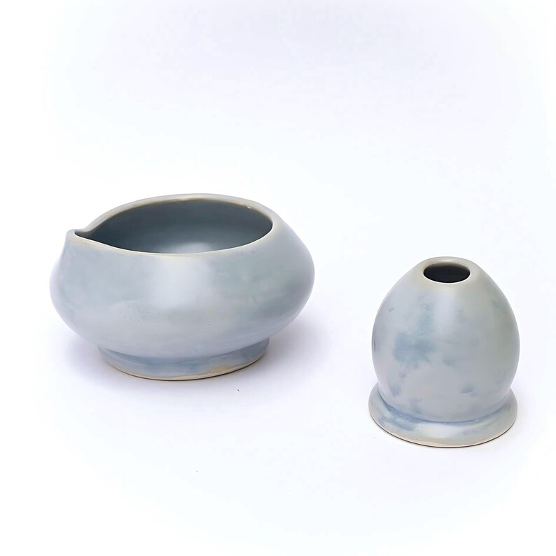 May include: Two light blue ceramic pieces on a white background. One is a small, rounded bowl with a spout. The other is a small, egg-shaped vase with a narrow opening at the top. Both pieces have a smooth, glossy finish.
