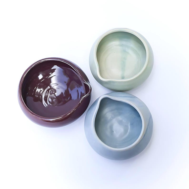 May include: Three ceramic bowls in shades of plum, seafoam green, and light blue. Each bowl has a rounded shape with a small spout on one side. The bowls are arranged in a triangular pattern against a white background.