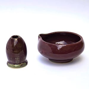 May include: A dark burgundy ceramic bowl and a small, matching vase. The bowl has a rounded shape with a spout, and the vase is egg-shaped with a green base. Both items have a glossy finish.