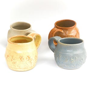 Könnte beinhalten: Vier handgefertigte Keramikbecher in verschiedenen Farben: Hellgelb, Beige, Orange und Blau. Jeder Becher hat eine abgerundete Form, einen großen Henkel und ein dekoratives geprägtes Blumenmuster. Die Becher stehen vor einem weißen Hintergrund.
