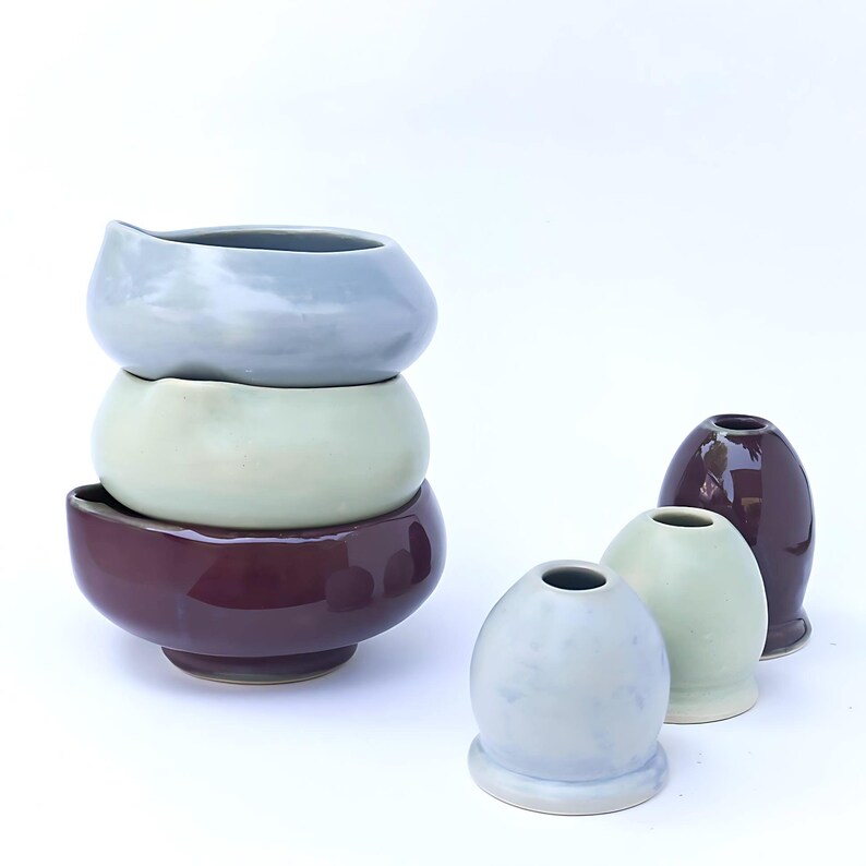 May include: A stack of three ceramic bowls in shades of gray, green, and burgundy. Beside them are three small, egg-shaped vases and one taller vase in similar colors. The pottery has a glossy finish and a minimalist design.
