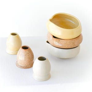 May include: A collection of handmade ceramic bowls and vases in various sizes and colors. The bowls are stacked, and the vases are arranged in front. The colors include cream, tan, and speckled brown. The objects have a smooth, matte finish.