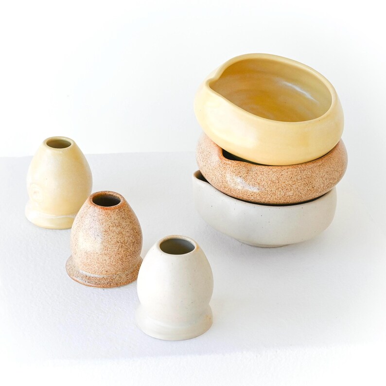 May include: A collection of handmade ceramic bowls and vases in various sizes and colors. The bowls are stacked, and the vases are arranged in front. The colors include cream, tan, and speckled brown. The objects have a smooth, matte finish.