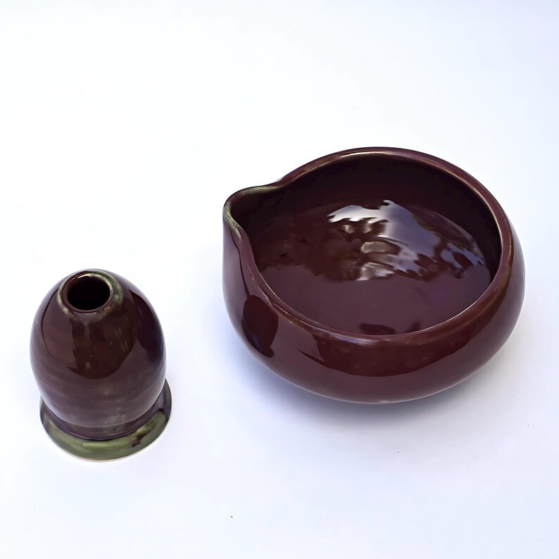 May include: A dark burgundy ceramic bowl with a spout and a small, matching vase. The vase is egg-shaped with a green base. Both items have a glossy finish and sit on a white surface.