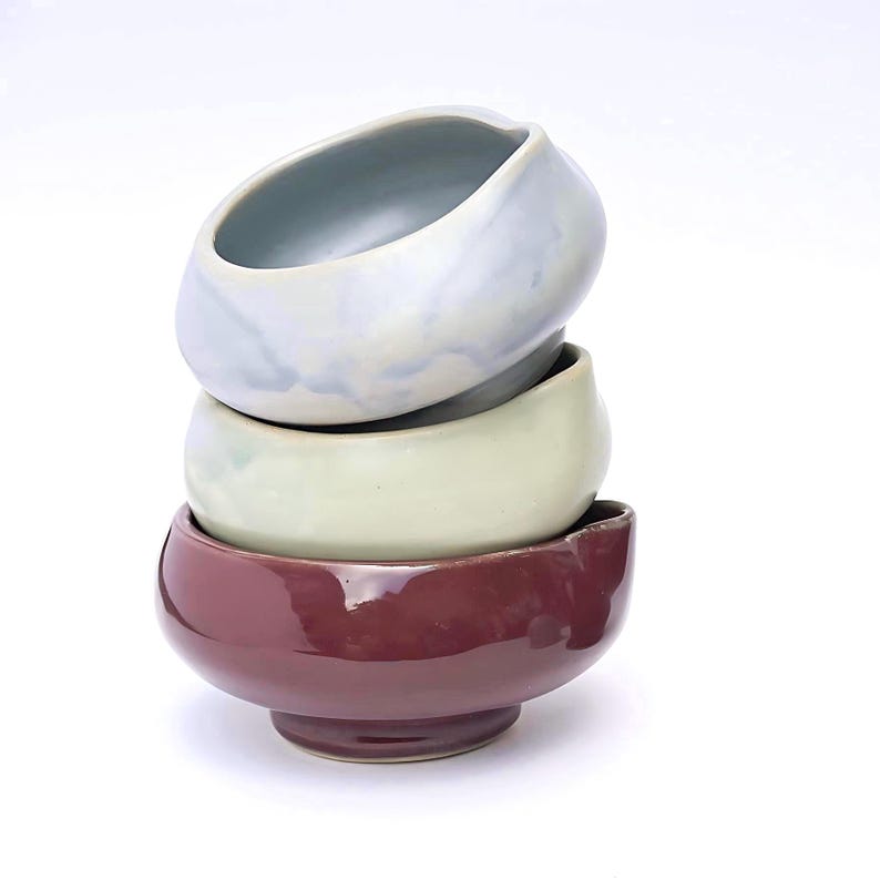 May include: A stack of four ceramic bowls in varying colors: pale blue, light green, and deep burgundy. The bowls have a smooth, glossy finish and a rounded shape, with a slightly asymmetrical design. The bowls are stacked on top of each other.