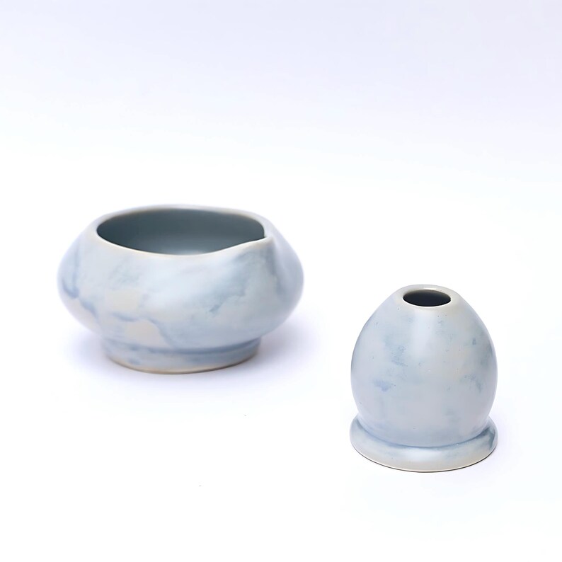 May include: Two light blue ceramic pieces. One is a small, rounded bowl with an irregular rim. The other is a small, egg-shaped vase with a narrow opening at the top. Both have a marbled appearance.