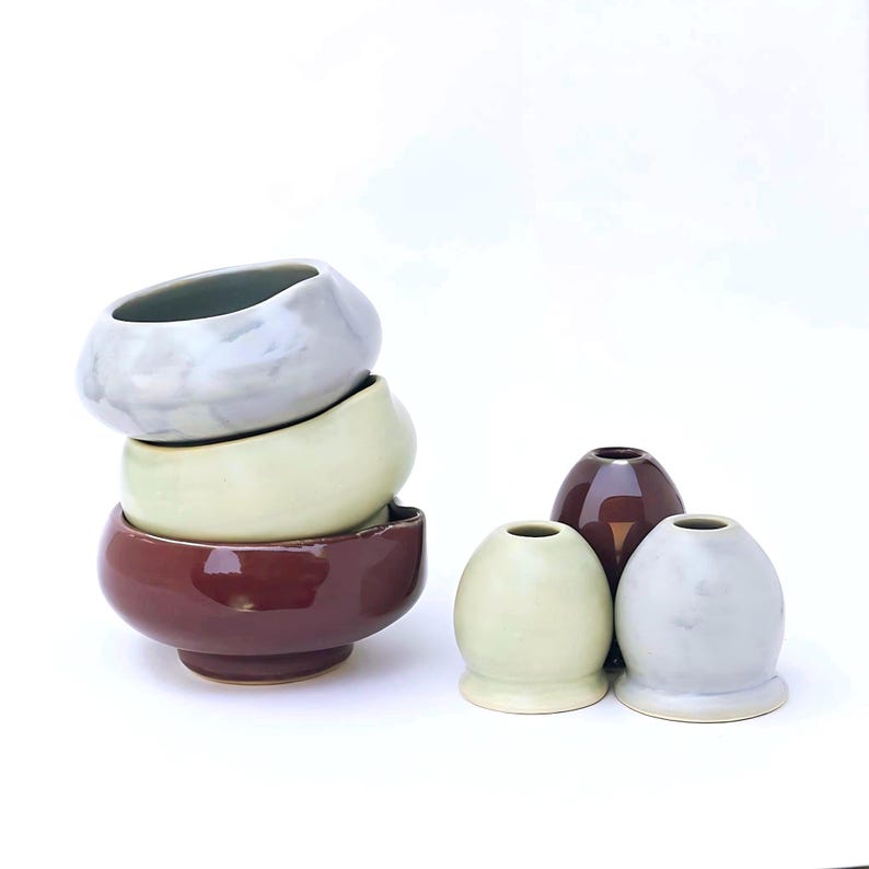 May include: A collection of ceramic bowls and vases in various colors. The bowls are stacked, with a deep burgundy bowl at the base, a pale green bowl in the middle, and a marbled gray bowl at the top. Three egg-shaped vases are also present.