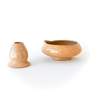 May include: Two ceramic pieces with a speckled, tan finish. One is a small, vase-like object with a flared base. The other is a rounded bowl with a spout. Both are on a white background.