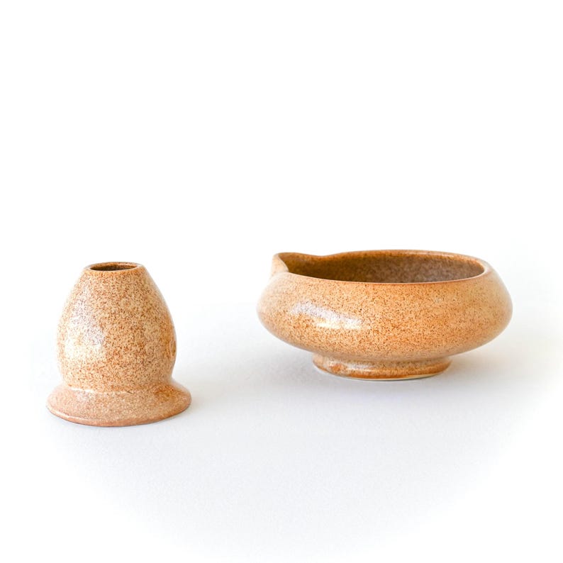 May include: Two ceramic pieces with a speckled, tan finish. One is a small, vase-like object with a flared base. The other is a rounded bowl with a spout. Both are on a white background.