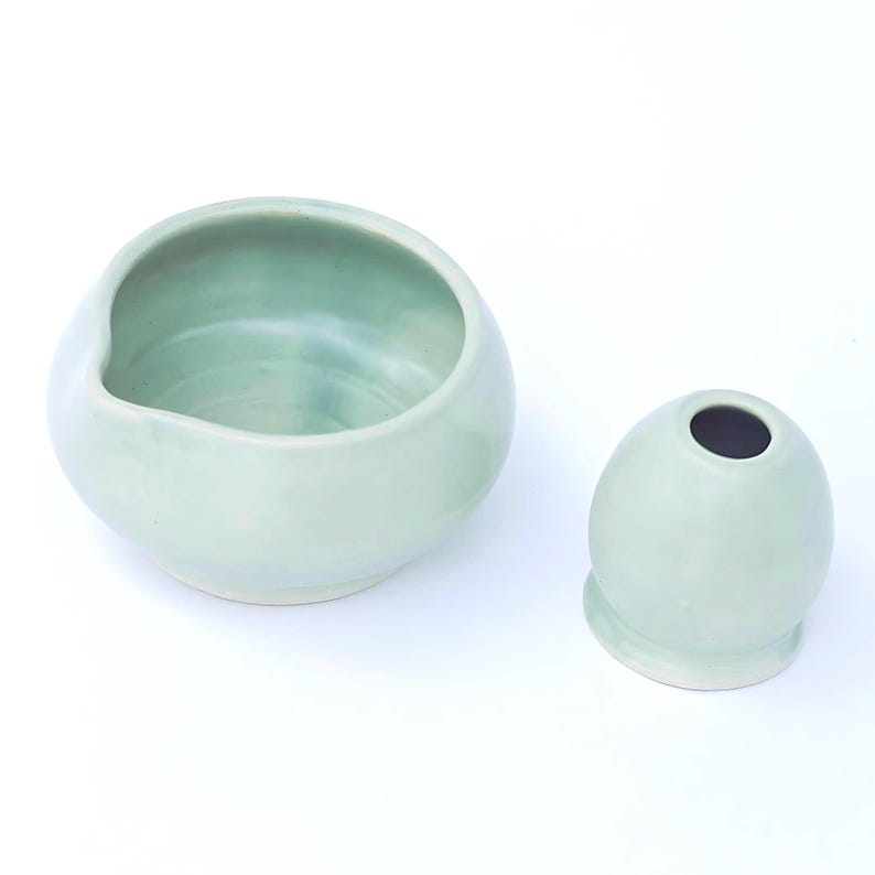 May include: Two ceramic pieces in a soft, pale green hue. One is a rounded bowl with a curved rim, and the other is a small, egg-shaped vase with a dark interior. Both pieces have a smooth, glazed finish.