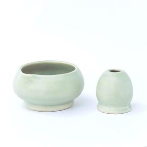 May include: Two ceramic pieces in a soft, pale green hue. One is a rounded bowl with a wide opening, and the other is a small, egg-shaped vase. Both have a smooth, glazed finish and sit on a white surface.