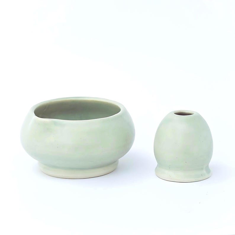 May include: Two ceramic pieces in a soft, pale green hue. One is a rounded bowl with a wide opening, and the other is a small, egg-shaped vase. Both have a smooth, glazed finish and sit on a white surface.