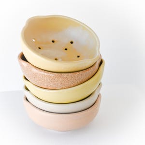 May include: A stack of six ceramic bowls in various pastel colors. The top bowl is a light yellow with several small holes. The other bowls are shades of brown, yellow, white, and pink. The bowls are stacked on top of each other.