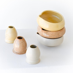 Pode incluir: Uma coleção de cerâmica artesanal em várias formas e cores. As peças incluem pequenos vasos em forma de ovo e uma pilha de três tigelas em tons de creme, castanho salpicado e amarelo. A cerâmica é exibida em uma superfície branca.