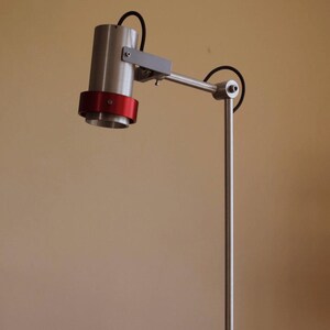May include: A silver desk lamp with a red accent ring on the shade. The lamp has a long, adjustable arm and a square, white base. A book titled "An Artist of the Floating World" by Kazuo Ishiguro is sitting on a wooden surface in front of the lamp.