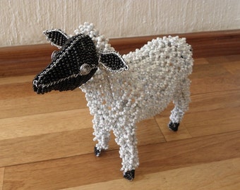 May include: A white beaded sheep with a black beaded head and legs. The sheep is standing on a wooden floor.
