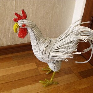 May include: A white beaded rooster with a red comb and yellow beak. The rooster is standing on a wooden floor.