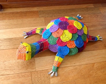 May include: A colorful beaded turtle sculpture with a rainbow pattern. The turtle is made of small, round beads in various colors, including red, orange, yellow, green, blue, purple, and white. The turtle's shell is a dome shape, and its legs and head are outstretched.