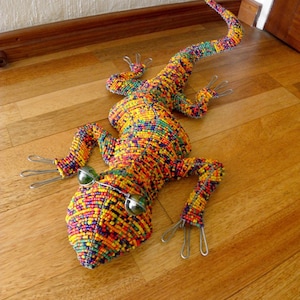 May include: A colorful beaded lizard figurine with green glass eyes. The lizard is made of small, colorful beads and is posed on a wooden floor.