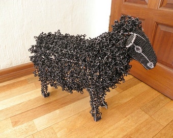May include: A black sheep sculpture made entirely of small, interconnected metal rings. The sculpture is standing on a wooden floor.