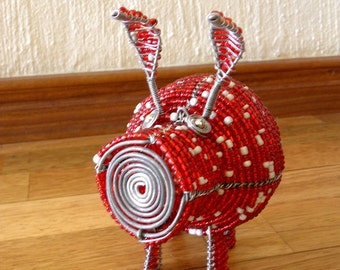 May include: A red and white beaded pig sculpture with wire legs and ears. The pig is standing on a wooden surface.