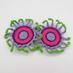 May include: Two handmade crochet appliques. Each applique features a central pink circle surrounded by a purple ring, with a lighter purple spiral in the center. Green, curved accents extend from the outer edge, creating a unique floral design.