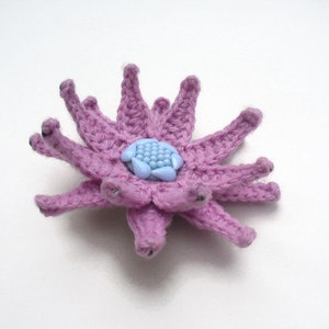 May include: A handmade crochet flower in shades of lavender and light blue. The flower has multiple petals and a central blue bead cluster. The crochet work is detailed, and the flower is set against a white background.