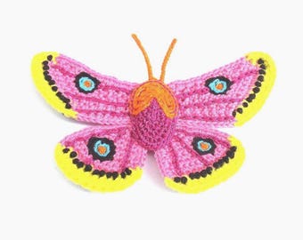 May include: A handmade crochet butterfly in shades of pink, yellow, and orange. The butterfly has pink wings with yellow edges and black dots. The body is a darker pink with orange antennae. The butterfly has blue and black eye details.