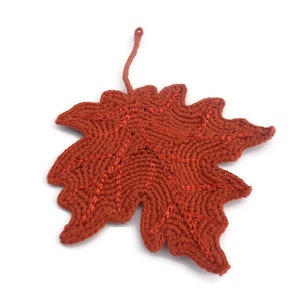 May include: A handcrafted, rust-colored crochet leaf with a stem. The leaf features detailed stitching in a darker shade of orange, creating a textured appearance. The leaf is likely a decorative item, possibly a coaster or a seasonal ornament.