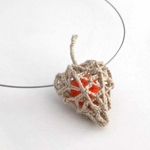 May include: A delicate necklace with a crocheted pendant featuring a small orange sphere. The pendant is shaped like a leaf and hangs from a silver chain.