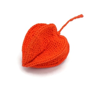 May include: A handmade, bright orange crocheted object resembling a Chinese lantern or a stylized fruit. The piece has a textured surface and a stem extending from the top. The item is set against a plain white background.