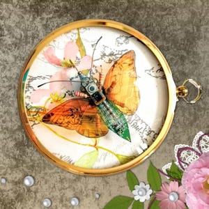 May include: A round, gold-framed artwork featuring a butterfly constructed from circuit board components. The butterfly has orange and yellow wings and is set against a backdrop of floral patterns and script. The frame includes a small loop for hanging.
