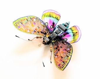 May include: A colorful butterfly sculpture with wings featuring circuit board patterns and rainbow hues. The body is composed of electronic components, with antennae and small details. The artwork is set against a white background.