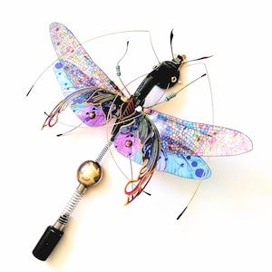 May include: A handcrafted dragonfly sculpture with iridescent wings in shades of purple, blue, and pink. The body is black with gold accents, and it features a silver ball and spring detail. The wings are made of a translucent material.