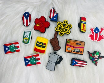 May include: A collection of 13 colorful shoe charms featuring Puerto Rican themes. The charms include a red hibiscus flower, a yellow can with the text "Coco Rico", a yellow can with the text "Cafe Bustelo", a brown mortar and pestle, a gray coffee pot, a brown box with the text "Sazon Goya", a red frog with a Puerto Rican flag, a yellow sun with a face, a red frog with a Puerto Rican flag, a red and blue Puerto Rican flag, a red and blue Puerto Rican flag, a red and blue Puerto Rican flag, and a red and blue Puerto Rican flag.