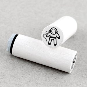 Op de afbeelding: Twee witte houten stempels met een zwart astronautenontwerp. Eén stempel staat rechtop en toont de astronautenillustratie, terwijl de andere horizontaal ligt. De stempels zijn cilindrisch met een grijs inktkussen.