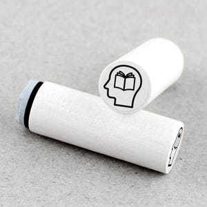 May include: White wooden stamp with a black outline of a head with an open book inside. The stamp is designed for creating custom art and crafts.