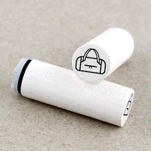 Op de afbeelding: Twee witte houten stempels met een zwarte omtrekillustratie van een handtas. Eén stempel staat rechtop en toont het handtassenontwerp, terwijl de andere horizontaal ligt en een zijaanzicht van hetzelfde ontwerp laat zien. De stempels liggen op een neutraal getinte ondergrond.
