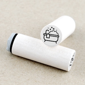 Peut inclure: Deux tampons en bois blancs avec des illustrations à l'encre noire. L'un des tampons représente une baignoire avec des bulles et une serviette. L'autre tampon a un design similaire. Les tampons sont cylindriques et ont une base en caoutchouc grise.