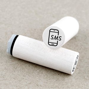 Könnte beinhalten: Zwei weiße Holzstempel mit schwarzen Tintenabdrücken. Ein Stempel zeigt ein Smartphone-Symbol mit den Buchstaben "SMS" darin. Der andere Stempel hat ein ähnliches Design. Die Stempel sind zylindrisch und haben eine hellfarbige Holzmaserung.